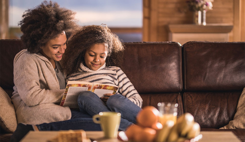 mother and daughter sit on couch reading together comfortably