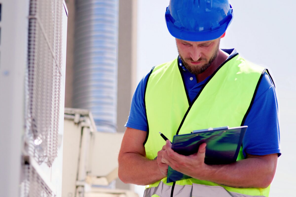 HVAC technician wearing hardhat, writing on a checklist on a clipboard with pen