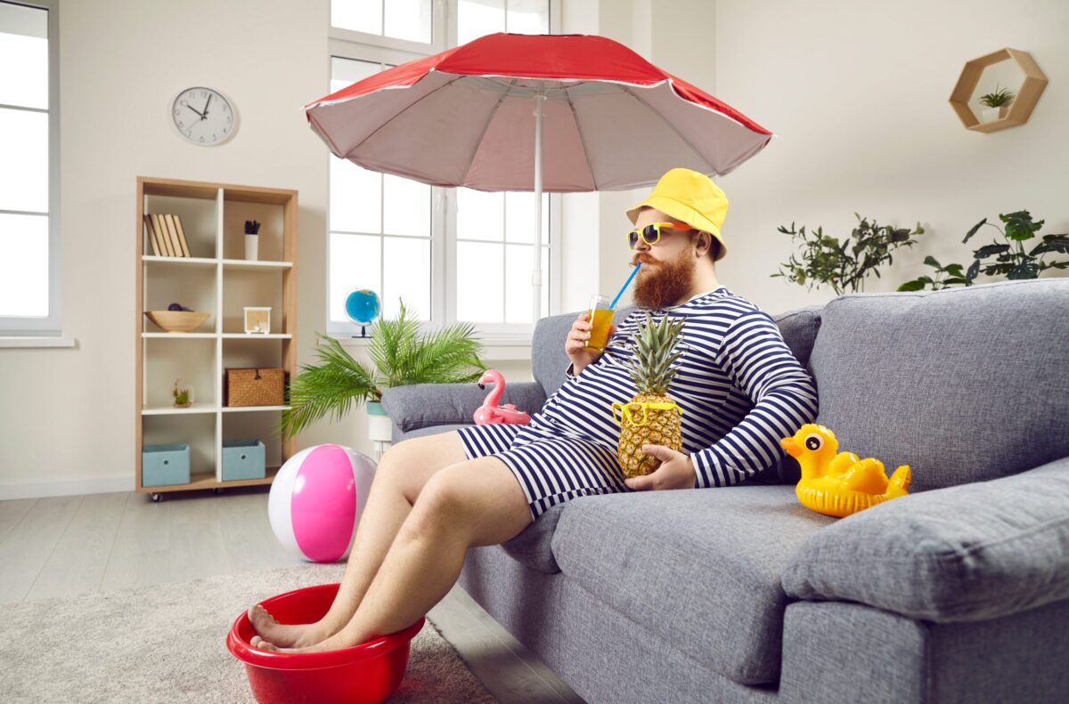 funny- man sitting on couch in beach wear with umbrella, rubber ducks, feet in bucket, pineapple,